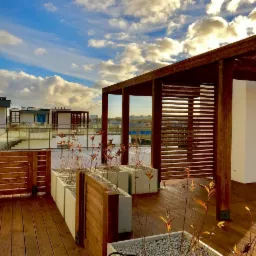 Drewniany taras na dachu budynku z konstrukcją pergoli, roślinami w donicach i widokiem na miasto w tle pod błękitnym niebem z chmurami.