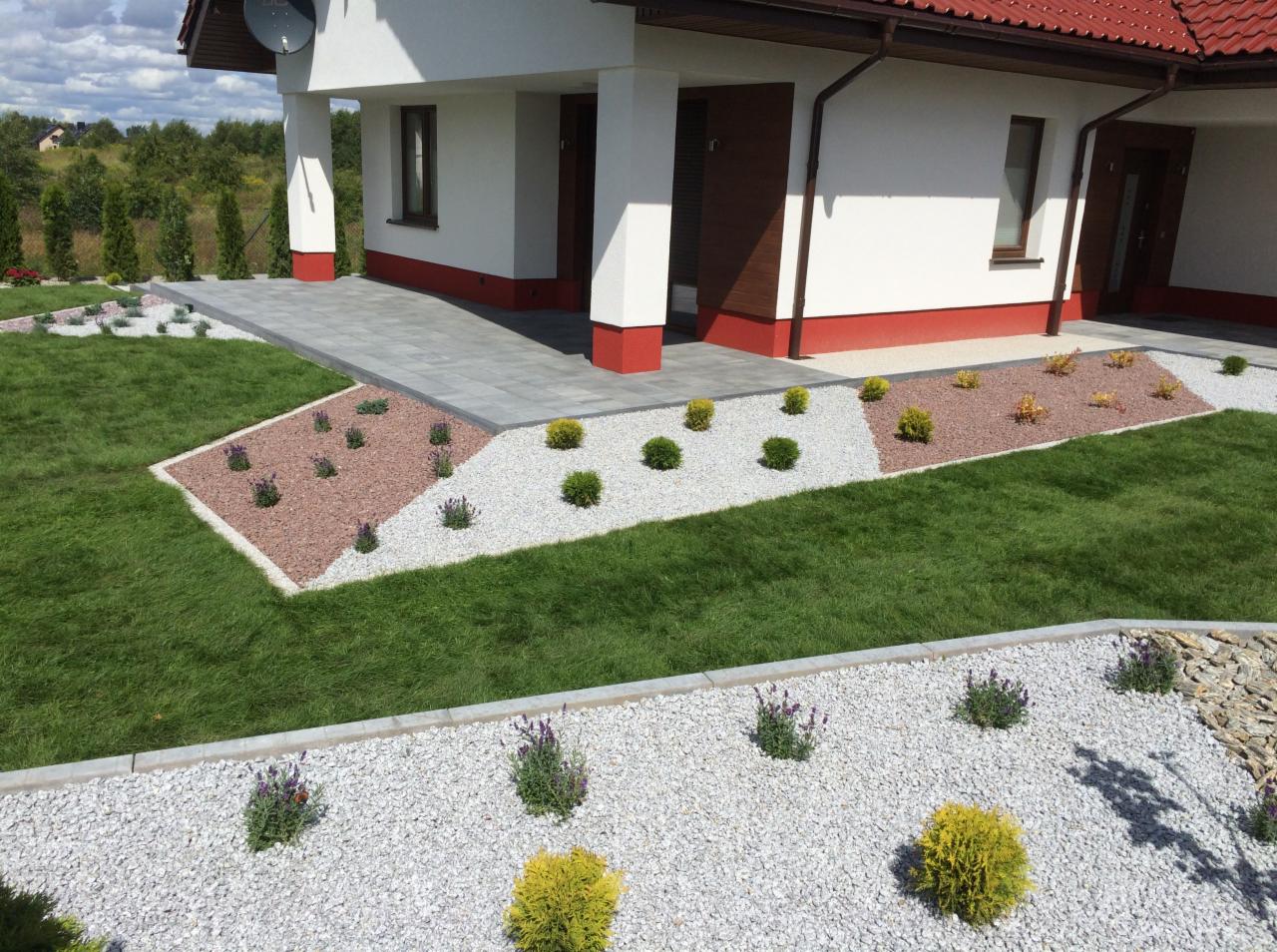 Nowoczesny ogród przydomowy z geometrycznymi rabatami wysypanymi żwirem, korą i obsadzonymi ozdobnymi krzewami oraz kostką brukową prowadzącą do wejścia.