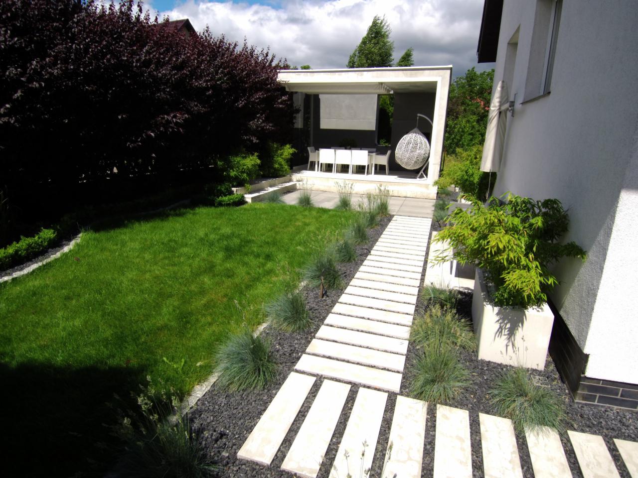 Nowoczesny ogród z równo przystrzyżonym trawnikiem, ścieżką z jasnych płyt na czarnym żwirze i altaną z białymi meblami.