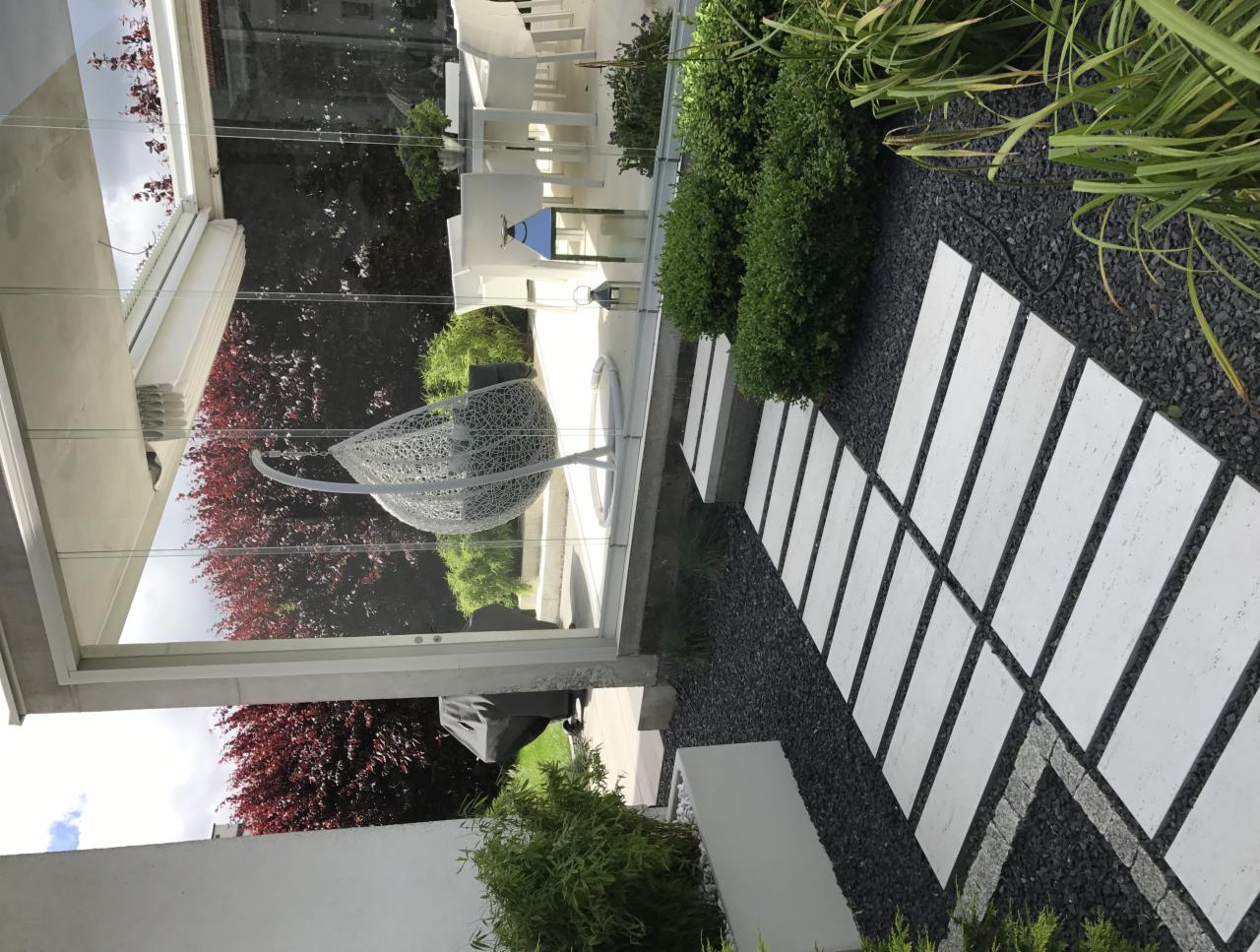 Nowoczesny ogród z ścieżką z białych płyt ułożonych w czarnym żwirze, obok szklanej ściany z widocznym ażurowym fotelem wiszącym i zielenią.