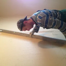 Pracownik w ubrudzonej odzieży roboczej, z poziomicą w dłoniach, sprawdza równość nałożonej gładzi na ścianie w trakcie remontu.