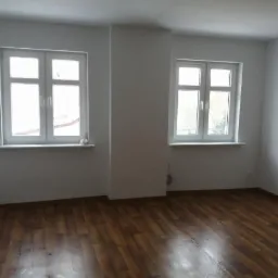 Pusty pokój po remoncie z brązowymi panelami podłogowymi, świeżo pomalowanymi białymi ścianami i dwoma oknami z białymi ramami.