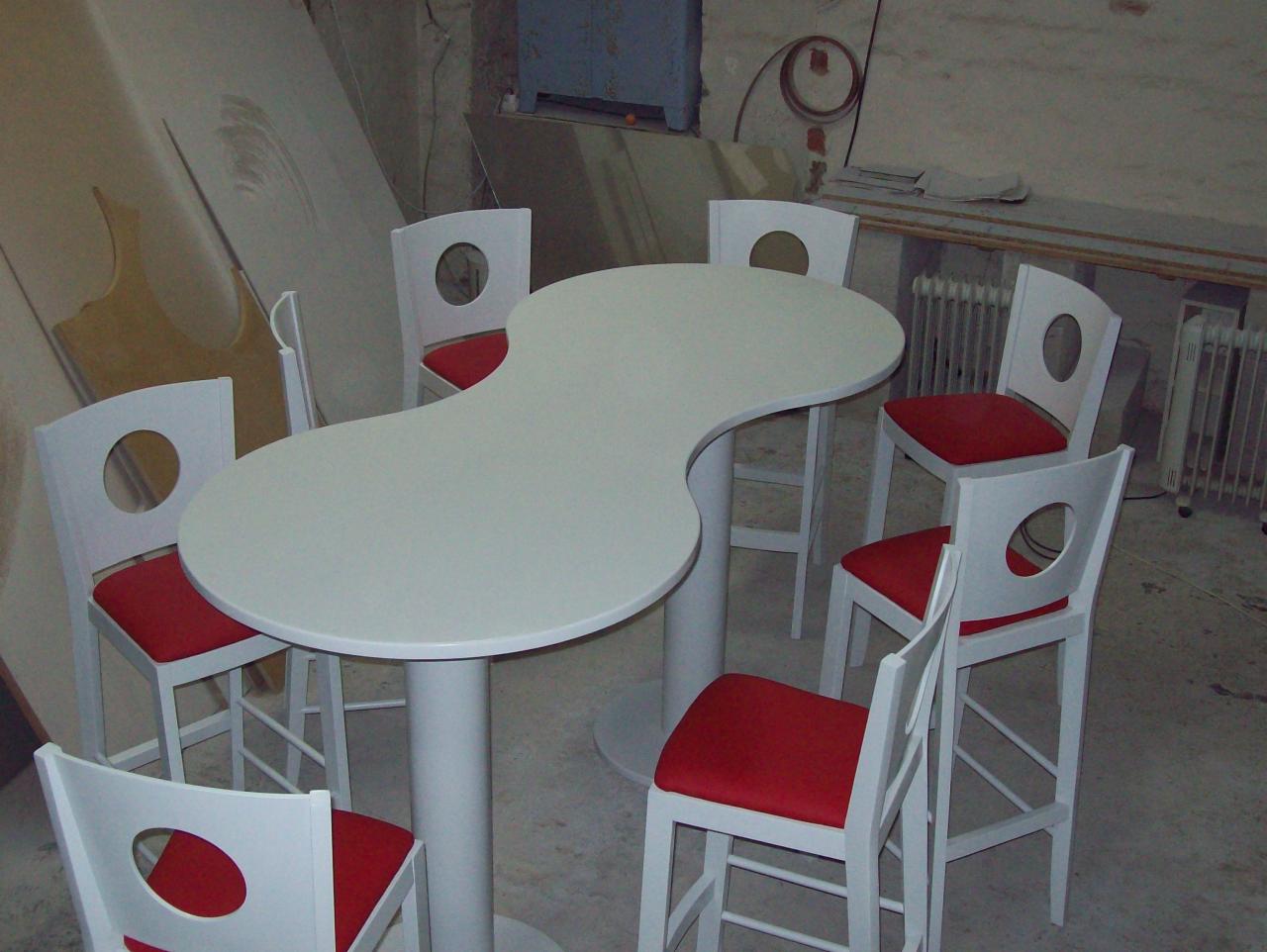 Biały stół o nieregularnym kształcie z sześcioma białymi krzesłami z czerwonymi siedziskami i okrągłymi otworami w oparciach, ustawiony w pomieszczeniu o surowym wykończeniu.