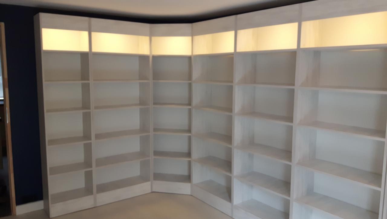 Białe, narożne półki z podświetleniem LED w górnej części, tworzące zabudowę w pokoju z ciemną ścianą.