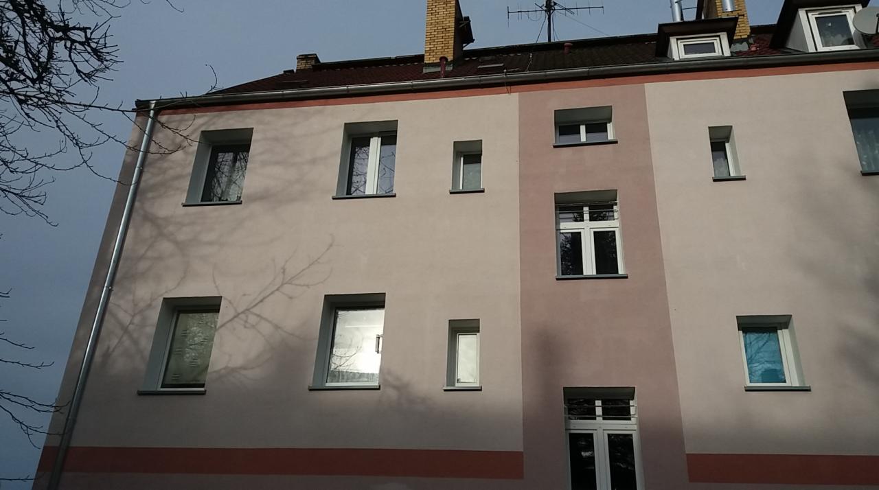 Elewacja budynku wielorodzinnego po renowacji, z widocznymi świeżo pomalowanymi fragmentami w odcieniach różu i beżu, okna z białymi ramami, na ścianie widoczne cienie drzew.