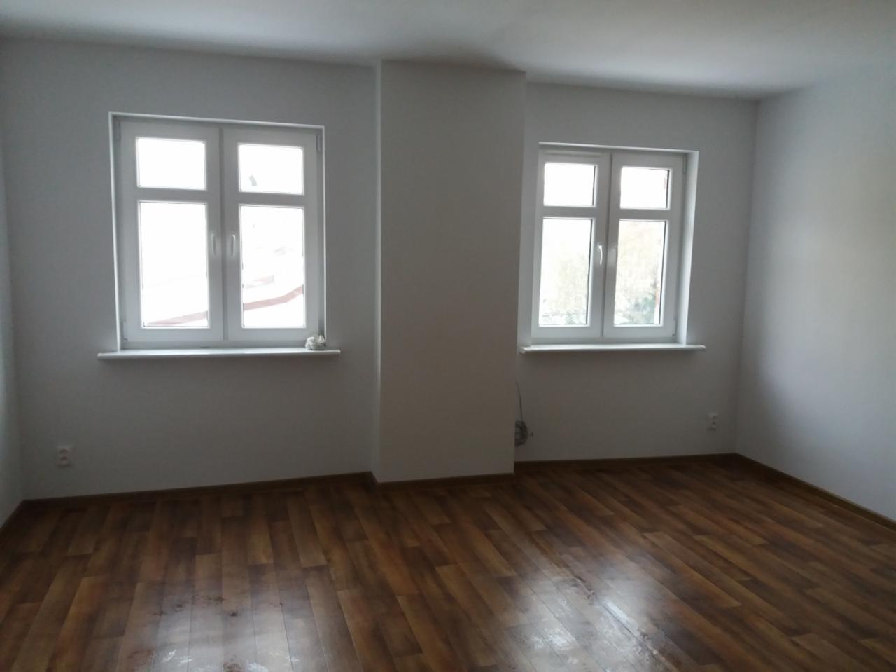 Pusty pokój po remoncie z brązowymi panelami podłogowymi, świeżo pomalowanymi białymi ścianami i dwoma oknami z białymi ramami.