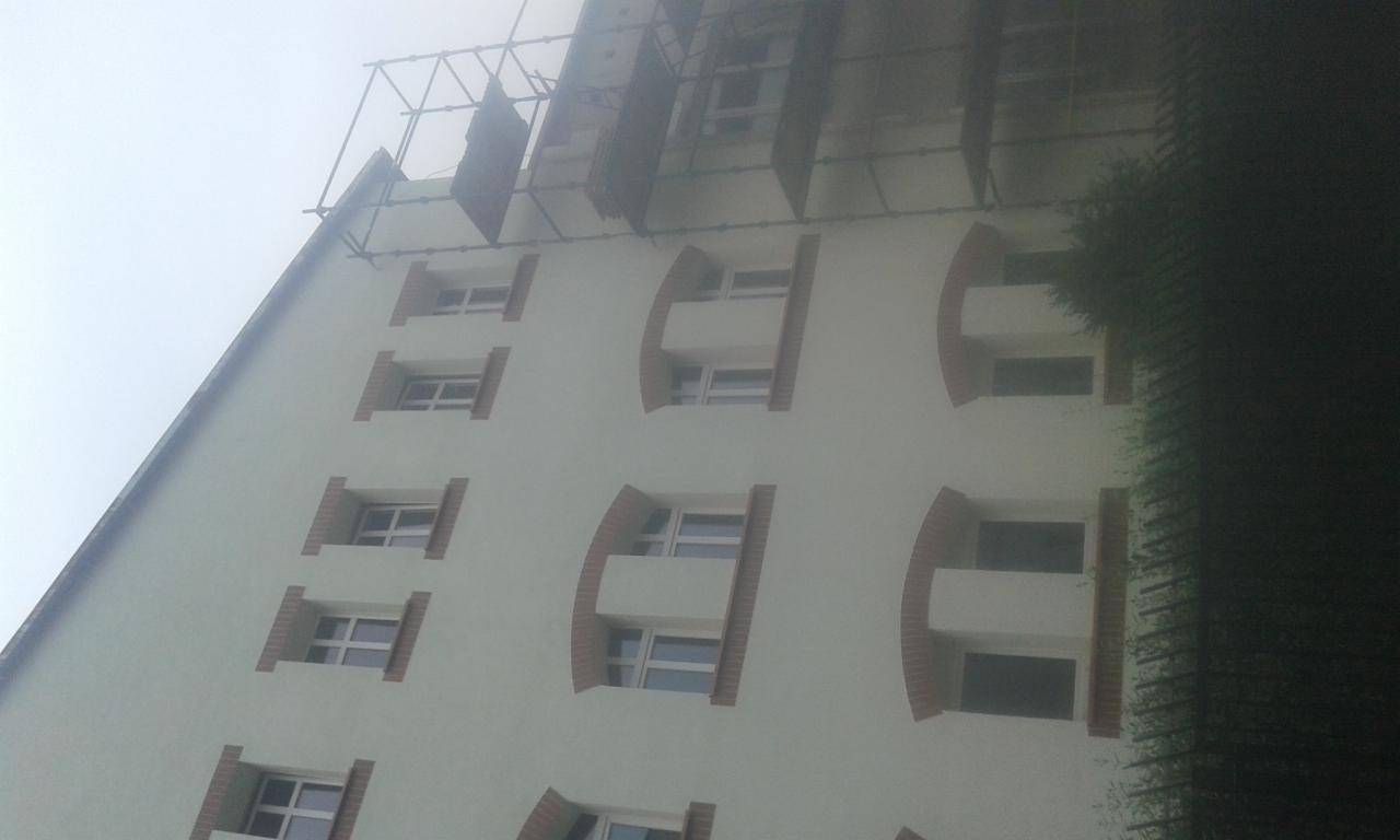 Elewacja budynku wielopiętrowego w trakcie prac remontowych, widoczne rusztowania i okna z dekoracyjnym obramowaniem, ujęcie z dołu w pochmurny dzień.