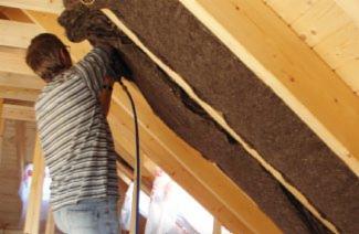 Pracownik w koszuli w paski instaluje brązową izolację z białym paskiem między krokwiami drewnianej konstrukcji dachu.