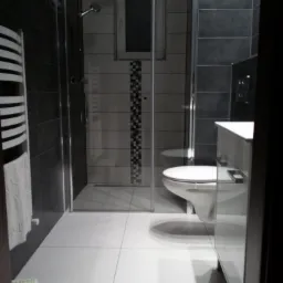 Nowoczesna łazienka z szarymi i białymi płytkami, kabiną prysznicową typu walk-in, wiszącą toaletą i grzejnikiem drabinkowym z ręcznikiem. Widoczny mały stołek dla dziecka.