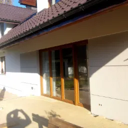 Ocieplona elewacja domu jednorodzinnego z brązowymi oknami przesuwnymi i ciemnobrązową dachówką ceramiczną, widok z boku na jasnym brukowanym tarasie.