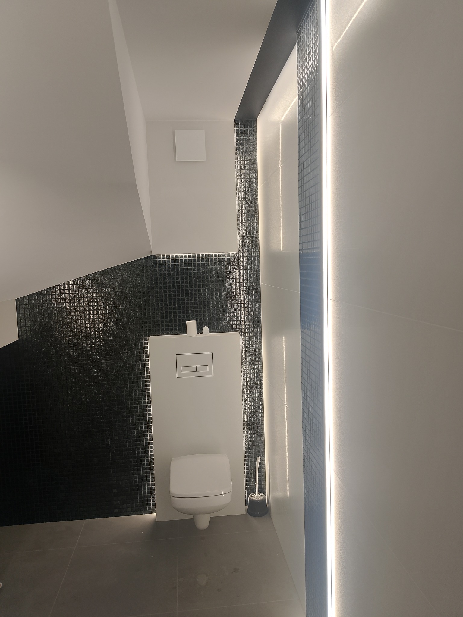 Nowoczesna łazienka z czarną mozaiką i podświetleniem LED. Wisząca toaleta z ukrytą spłuczką. Minimalistyczny design z szarymi płytkami podłogowymi.