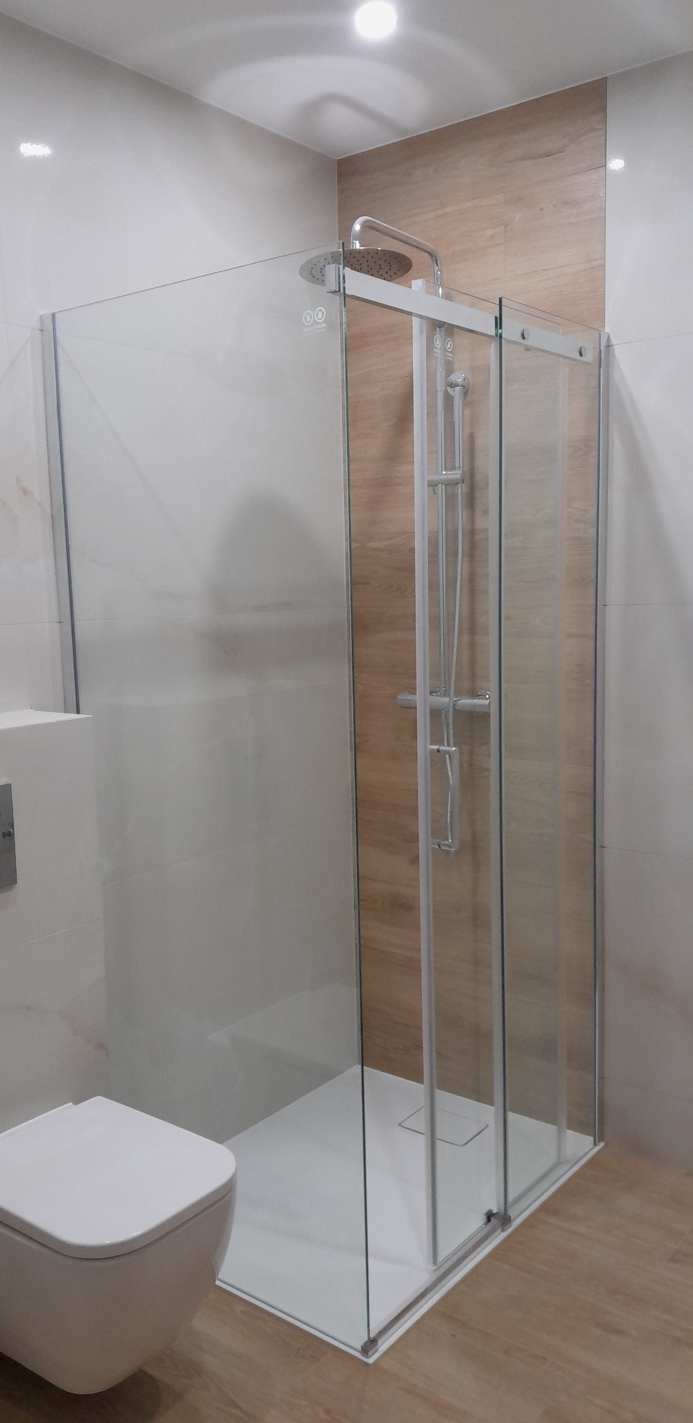 Nowoczesna kabina prysznicowa z przesuwnymi drzwiami, wykończona płytkami imitującymi drewno, z widoczną białą miską WC w minimalistycznym stylu.