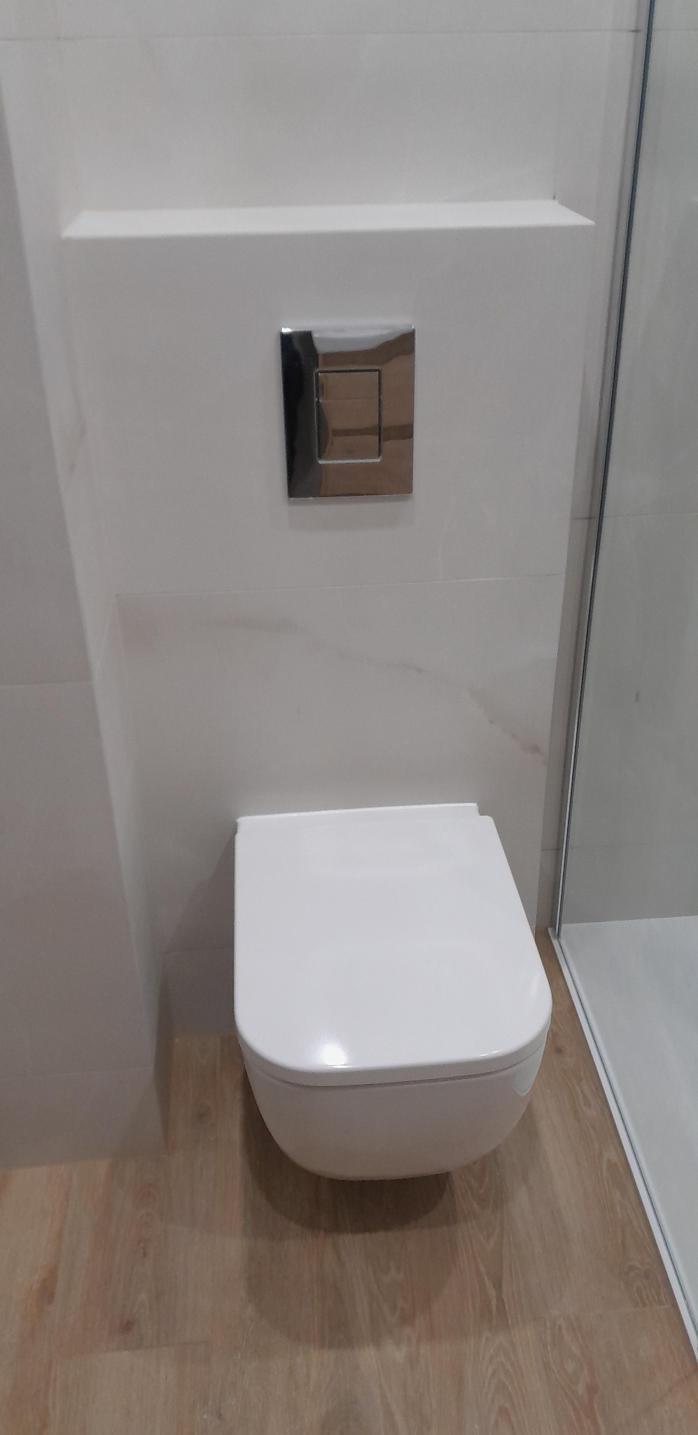 Biała, podwieszana toaleta z chromowanym przyciskiem spłukującym na tle marmurowych płytek i drewnopodobnej podłogi, widok z przodu, minimalistyczny design.