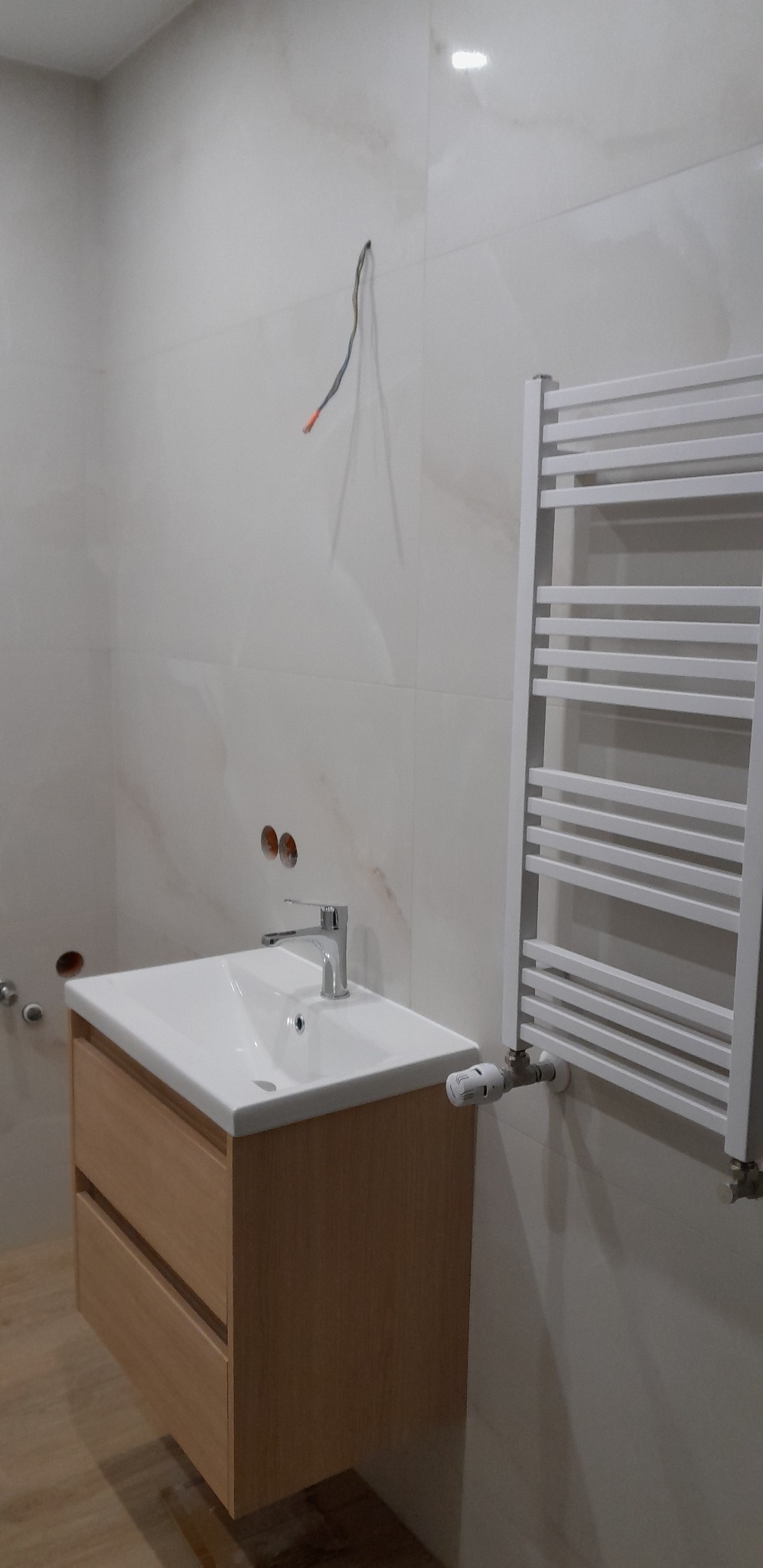 Jasna łazienka po remoncie: umywalka z szafką w odcieniach drewna, biały grzejnik drabinkowy, marmurowe płytki ścienne i wystające kable.