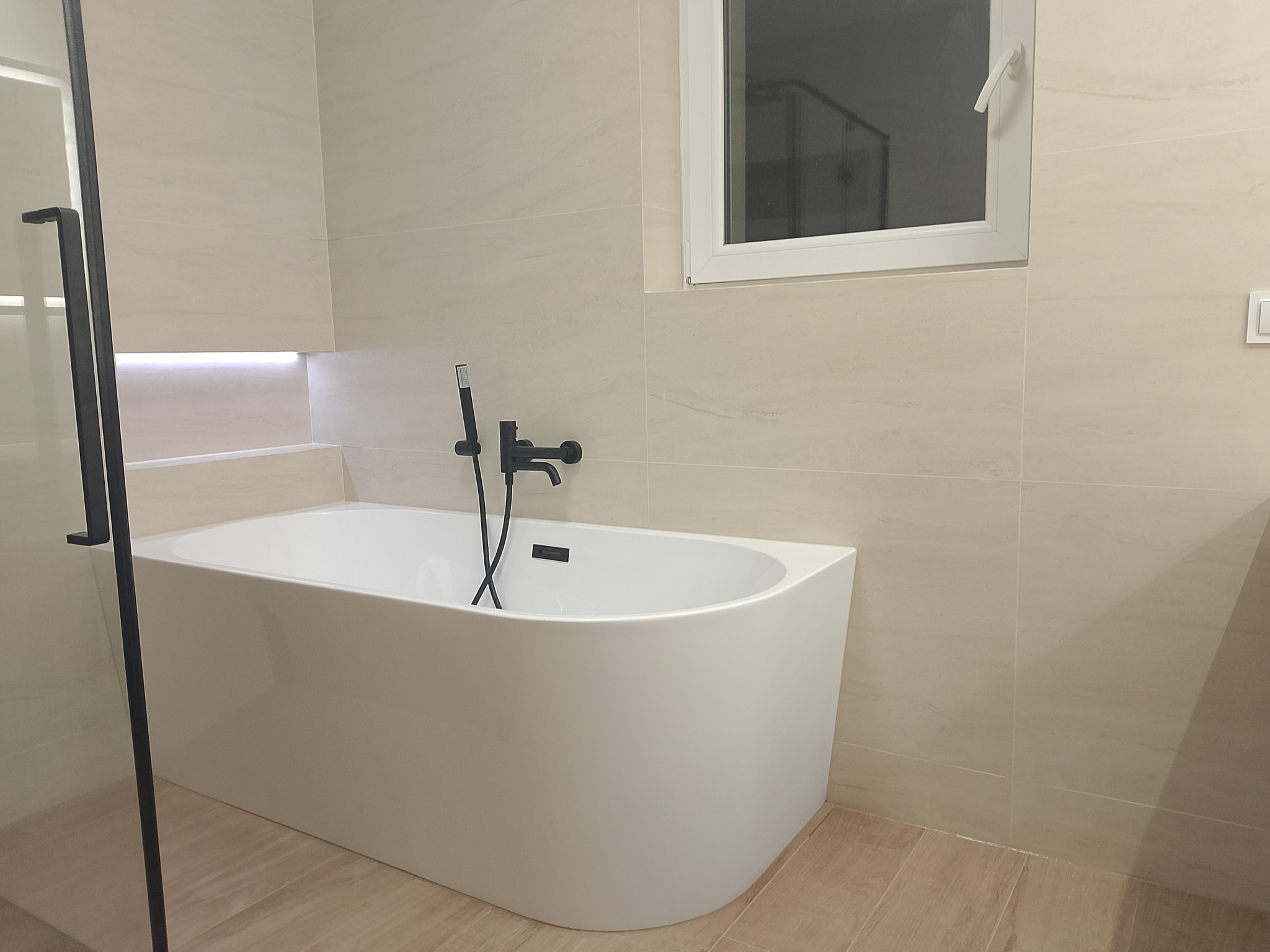 Nowoczesna łazienka z białą wanną wolnostojącą i czarną armaturą na tle beżowych płytek imitujących marmur. Minimalistyczny design, oświetlenie LED w niszy.