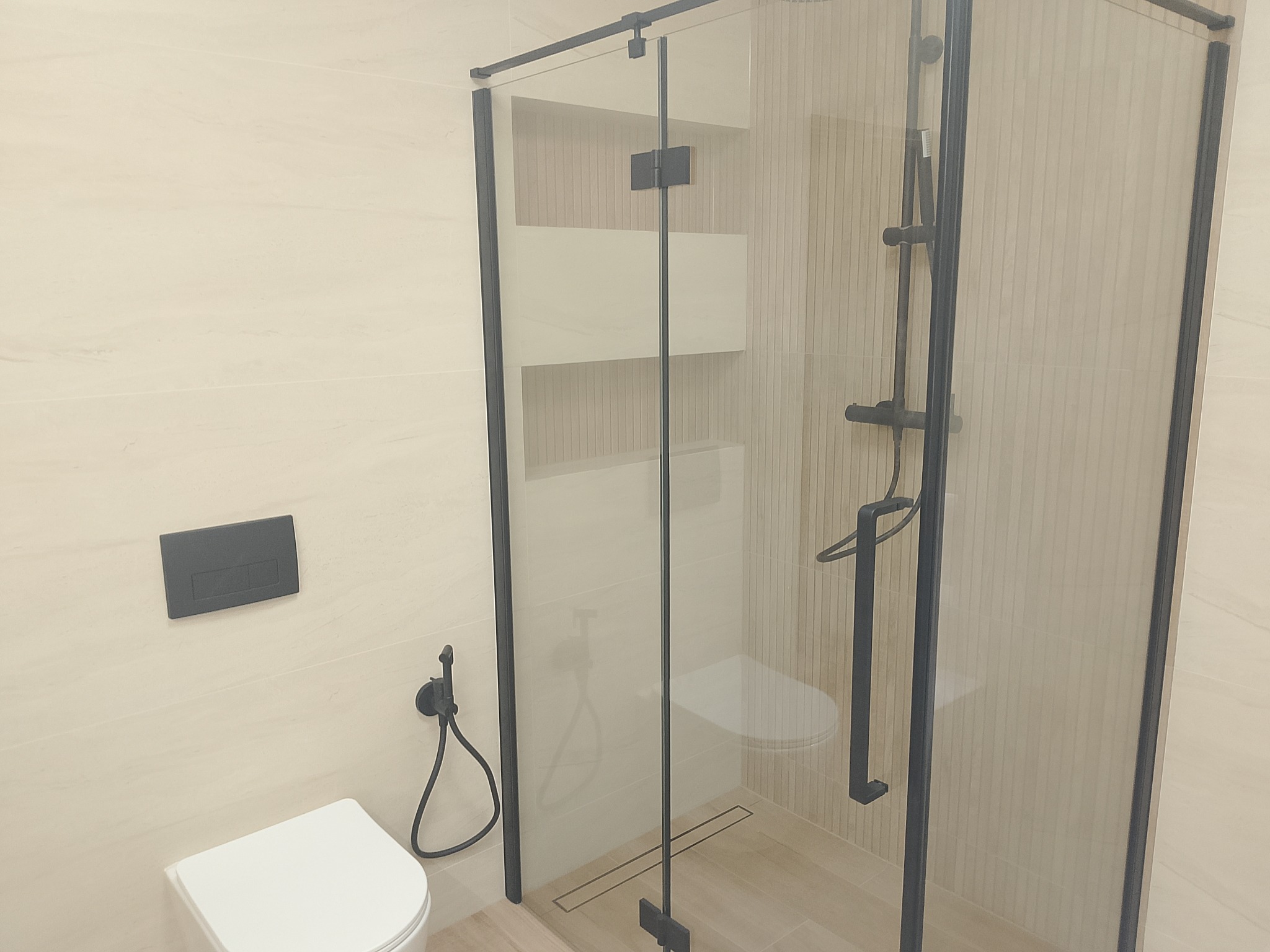 Nowoczesna łazienka z kabiną prysznicową typu walk-in z czarnymi profilami, podwieszaną toaletą i beżowymi płytkami. Minimalistyczny design.