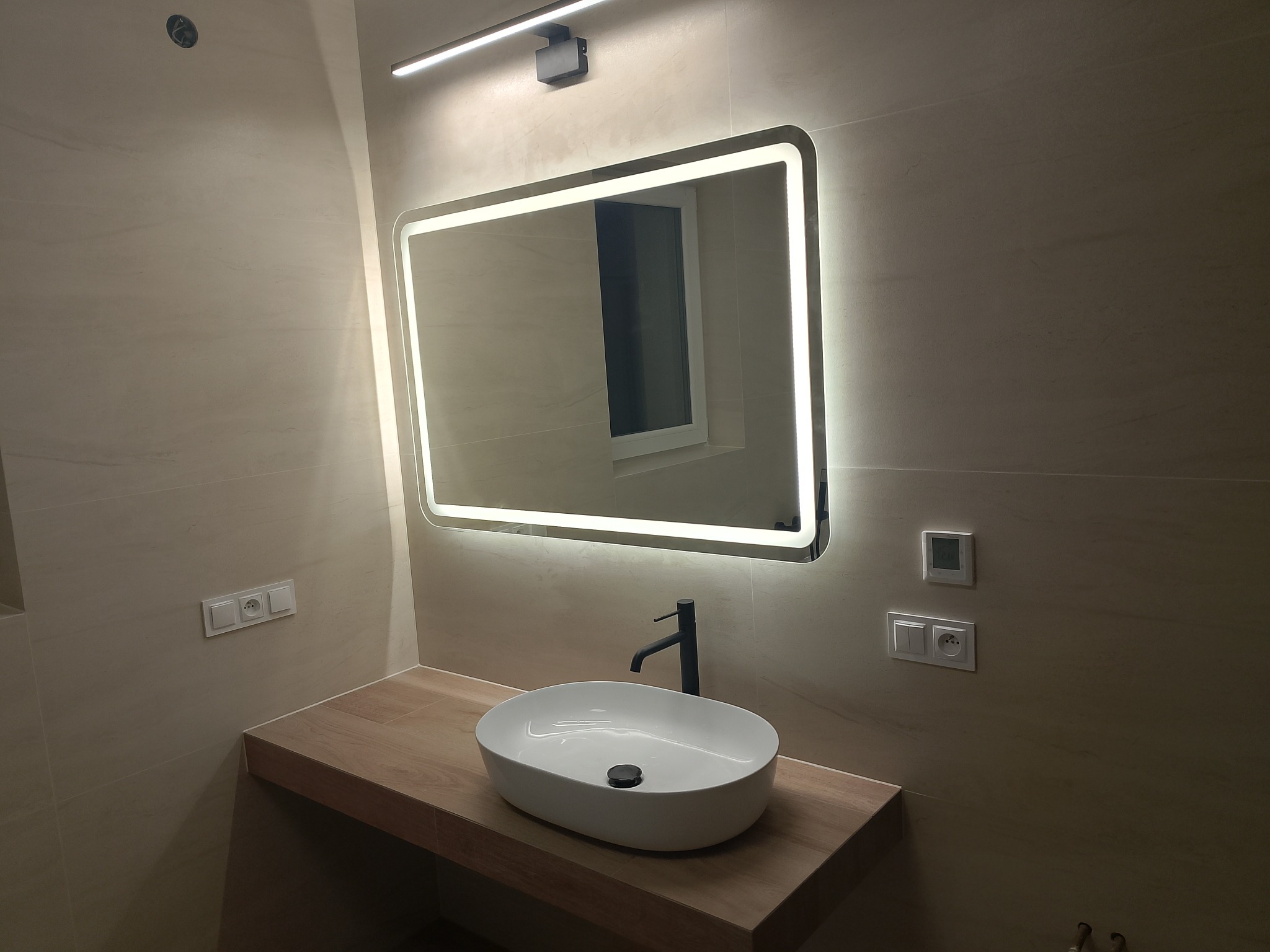 Nowoczesna łazienka z podświetlanym lustrem, owalną umywalką na drewnianym blacie i beżowymi płytkami. Minimalistyczny design, czarna bateria, gniazdka i włączniki na ścianie.