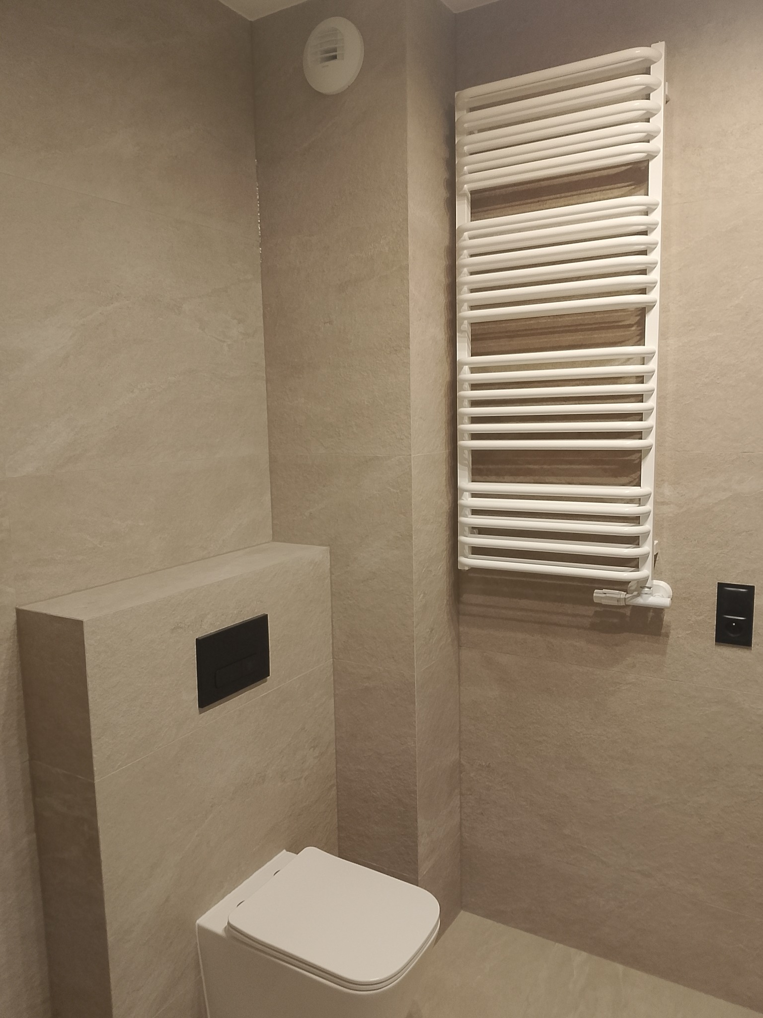 Beżowa łazienka po remoncie: wisząca toaleta z czarnym przyciskiem, grzejnik drabinkowy i płytki imitujące kamień. Minimalistyczny design, ciepłe oświetlenie.