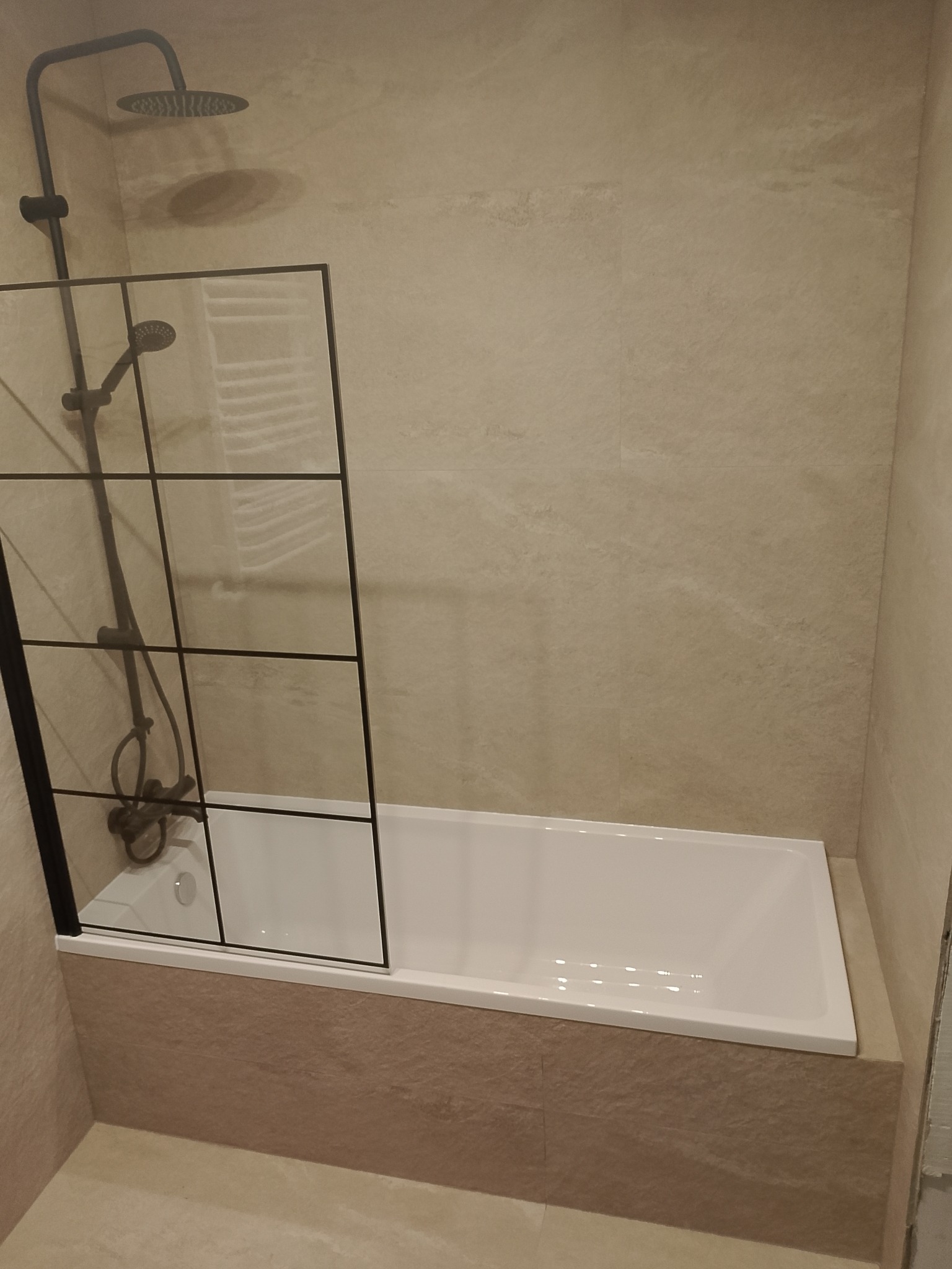 Nowoczesna łazienka z beżowymi płytkami, białą wanną i czarną, szklaną kabiną prysznicową. Minimalistyczny design, stonowana kolorystyka, widoczny fragment baterii prysznicowej.