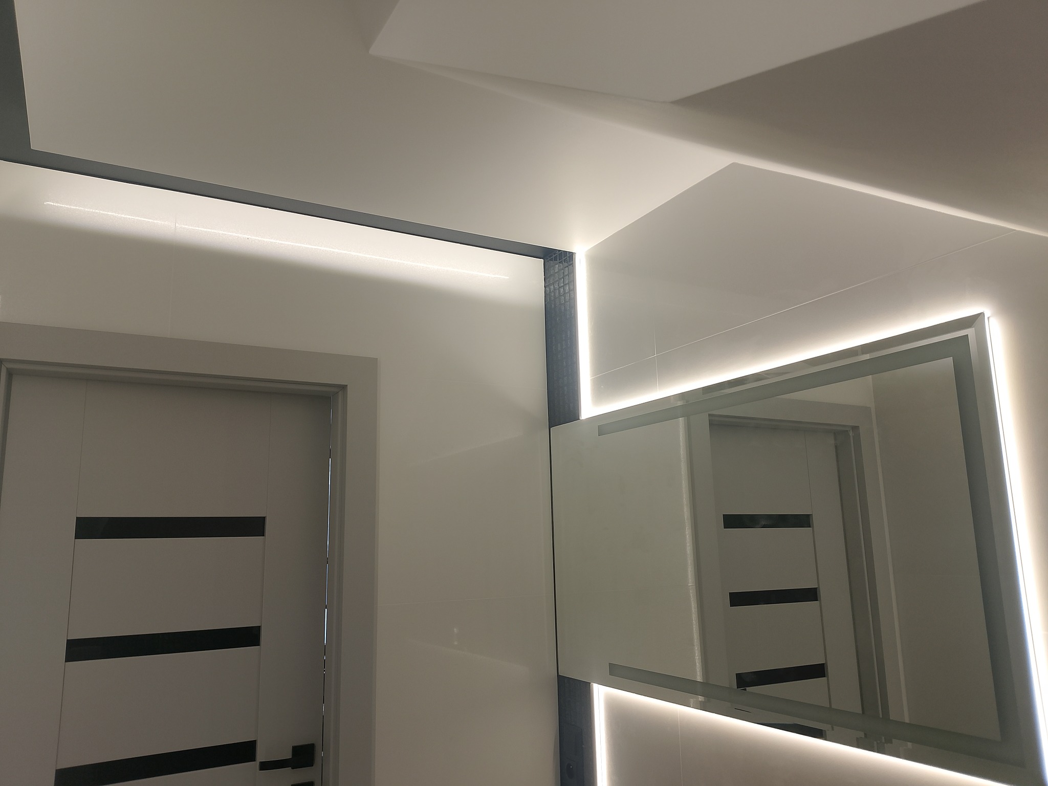 Nowoczesna łazienka z podświetlanym lustrem i drzwiami z czarnymi akcentami. Białe płytki na ścianach, minimalistyczny design, oświetlenie LED.