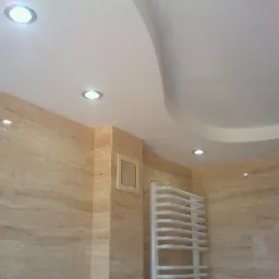 Wykończona łazienka z beżowymi płytkami imitującymi trawertyn, podwieszany sufit z wpuszczonym oświetleniem LED i białym grzejnikiem drabinkowym.