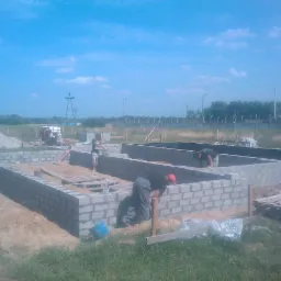 Budowa fundamentów domu z bloczków betonowych, widoczni pracownicy w trakcie murowania, w tle krajobraz z wieżą kościelną.