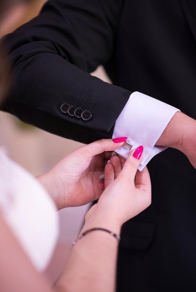 Kobieta zapina spinki do mankietów w czarnym garniturze mężczyzny, widoczne dłonie z różowym manicure.