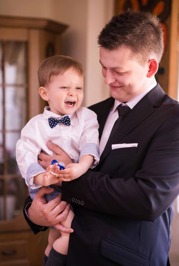 Uśmiechnięty mężczyzna w garniturze trzyma na rękach roześmianego chłopca w koszuli i muszce w kropki.