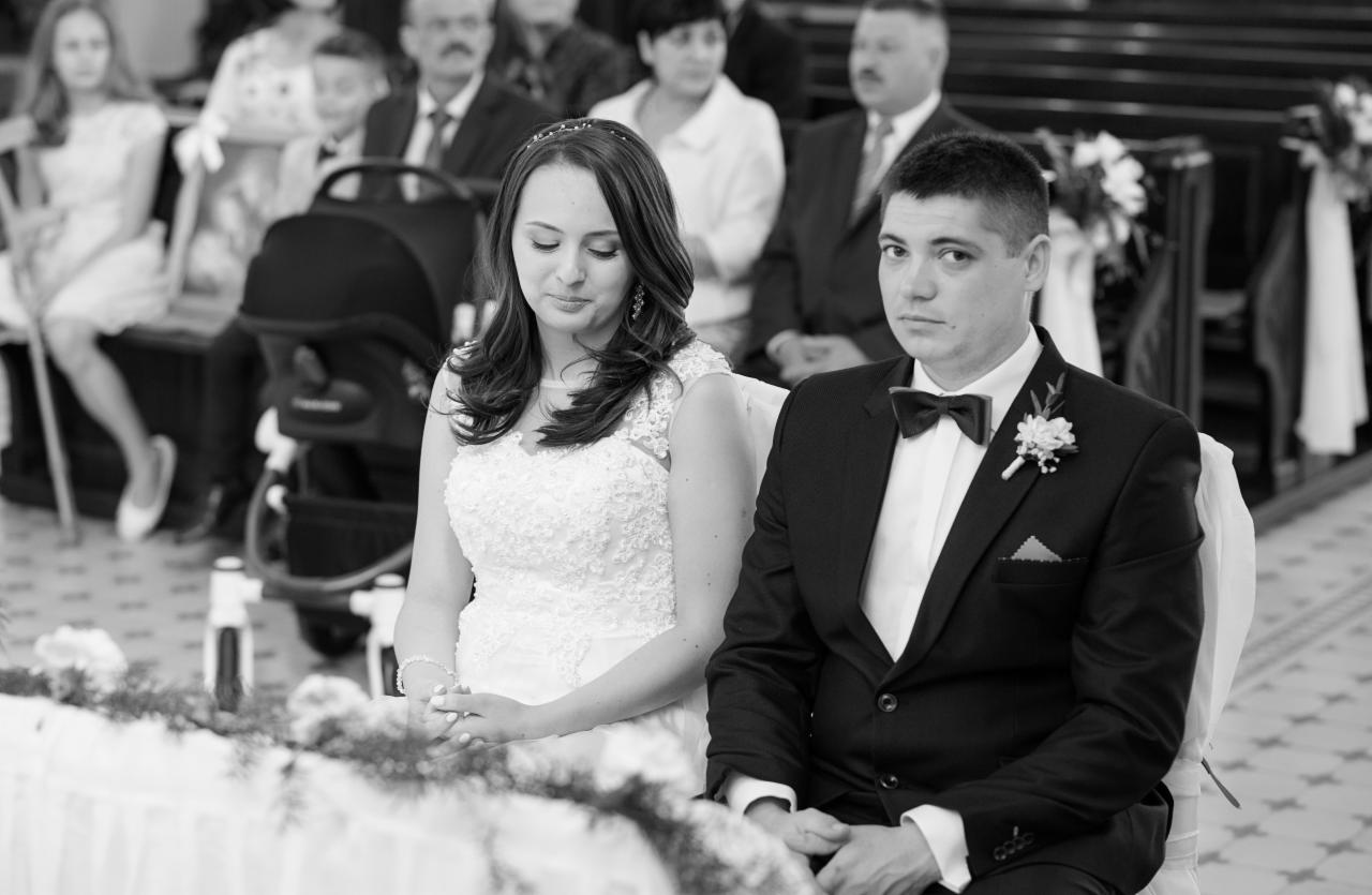 Czarno-białe zdjęcie pary młodej siedzącej w kościele podczas ceremonii ślubnej, z gośćmi w tle i wózkiem dziecięcym po lewej stronie.