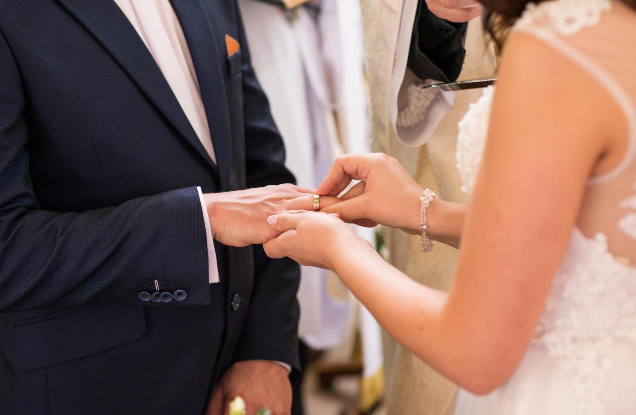 Panna młoda zakłada obrączkę na palec pana młodego podczas ceremonii ślubnej w kościele, widoczny fragment granatowego garnituru z pomarańczową poszetką.