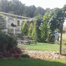 Zadbany ogród z trawnikiem, drzewem o zwisających gałęziach, kamienną rabatą, leżakami i białą pergolą z roślinami w donicach.