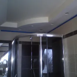 Nowoczesna kabina prysznicowa z chromowanymi profilami, widok na ułożone płytki w odcieniach beżu i brązu, sufit z podwieszanym elementem i oświetleniem punktowym, zabezpieczony niebieską taśmą...