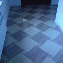 Ułożona pod kątem podłoga z kwadratowych płytek w dwóch odcieniach, z dekoracyjnym wzorem w narożnikach, radiator w tle, widok z góry.