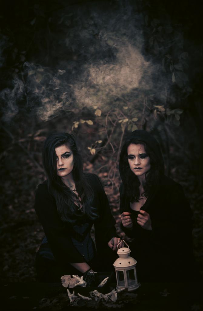 Dwie kobiety w ciemnych strojach i makijażu w stylu gotyckim siedzą w lesie z białą latarnią i kośćmi zwierząt, otoczone mgłą.
