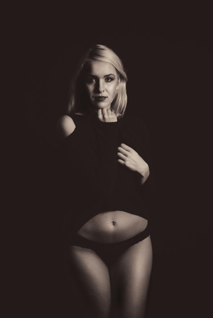 Portret kobiety w czarnym swetrze z odkrytym ramieniem, pozującej na ciemnym tle, z dłonią pod brodą i drugą na brzuchu.