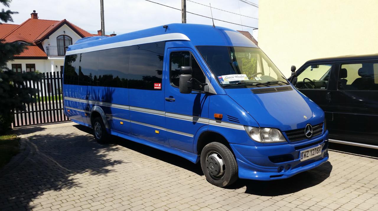Niebieski bus Mercedes-Benz Sprinter z napisem 'Goście Weselni' na przedniej szybie, zaparkowany na brukowanym podjeździe przed domem z czerwoną dachówką.