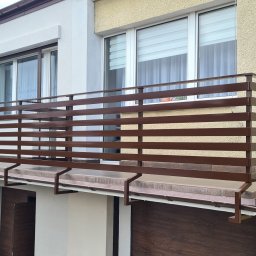 On Yacht - Balkon z poziomą, brązową balustradą metalową, zamontowaną na dwóch budynkach o różnej kolorystyce elewacji: białej i żółtej.