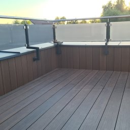 On Yacht - Nowoczesny balkon z brązową podłogą z desek kompozytowych i balustradą ze stali nierdzewnej z panelami z matowego szkła, mocowaną za pomocą czarnych uchwytów.