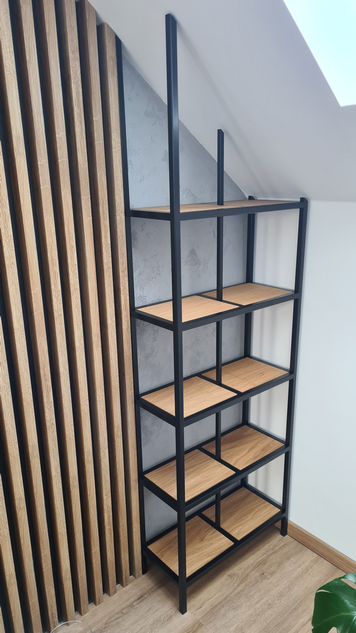 Czarny, metalowy regał z drewnianymi półkami, wykonany w Osielsku, ustawiony na poddaszu, na tle drewnianej ściany z pionowych listew i szarej ściany ze skosem.