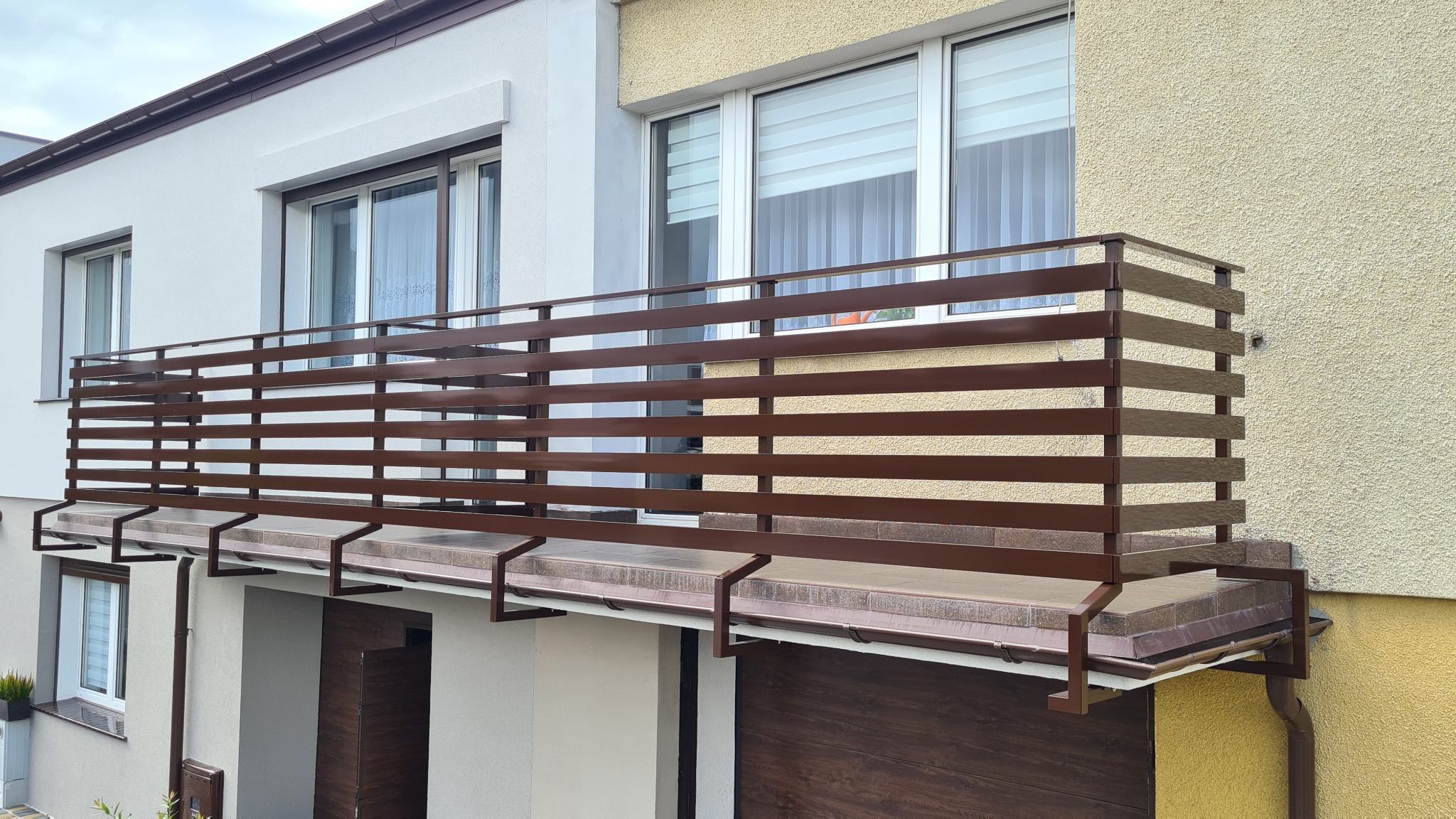Balkon z poziomą, brązową balustradą metalową, zamontowaną na dwóch budynkach o różnej kolorystyce elewacji: białej i żółtej.
