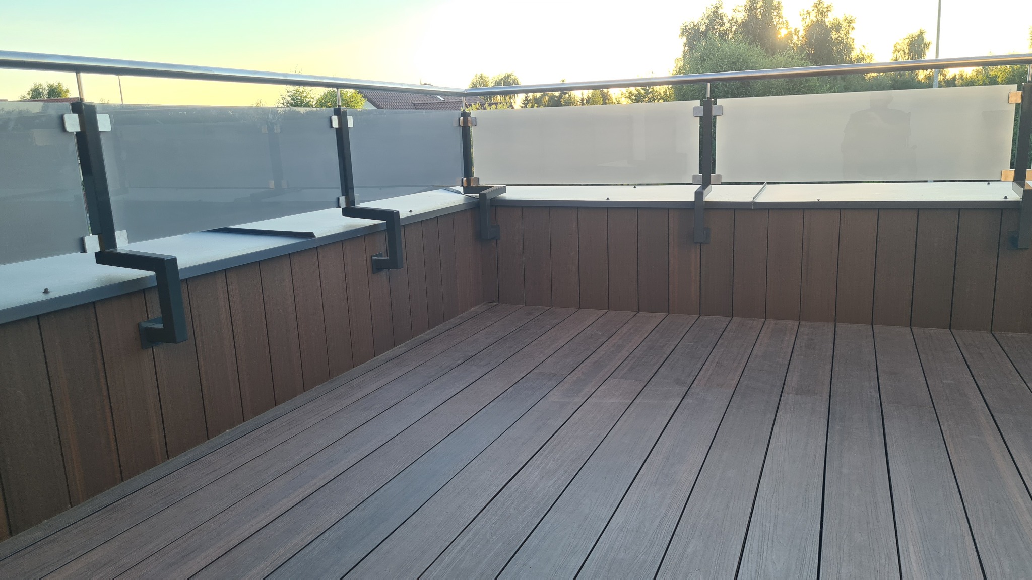 Nowoczesny balkon z brązową podłogą z desek kompozytowych i balustradą ze stali nierdzewnej z panelami z matowego szkła, mocowaną za pomocą czarnych uchwytów.