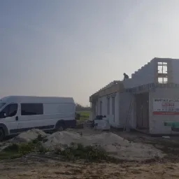 Budowa domu z białego bloczka: stan surowy otwarty, widoczny pracownik na dachu, biały samochód dostawczy firmy, sterta piasku na pierwszym planie, tablica informacyjna z nazwą firmy budowlanej...