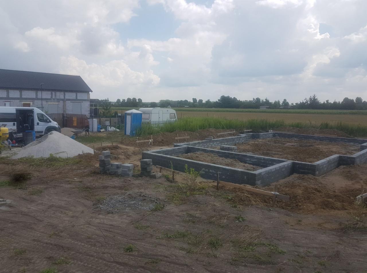 Budowa fundamentów pod dom: widok na wykop z częściowo wzniesionymi ścianami fundamentowymi z bloczków betonowych, w tle zaparkowany bus, przyczepa kempingowa i przenośna toaleta.