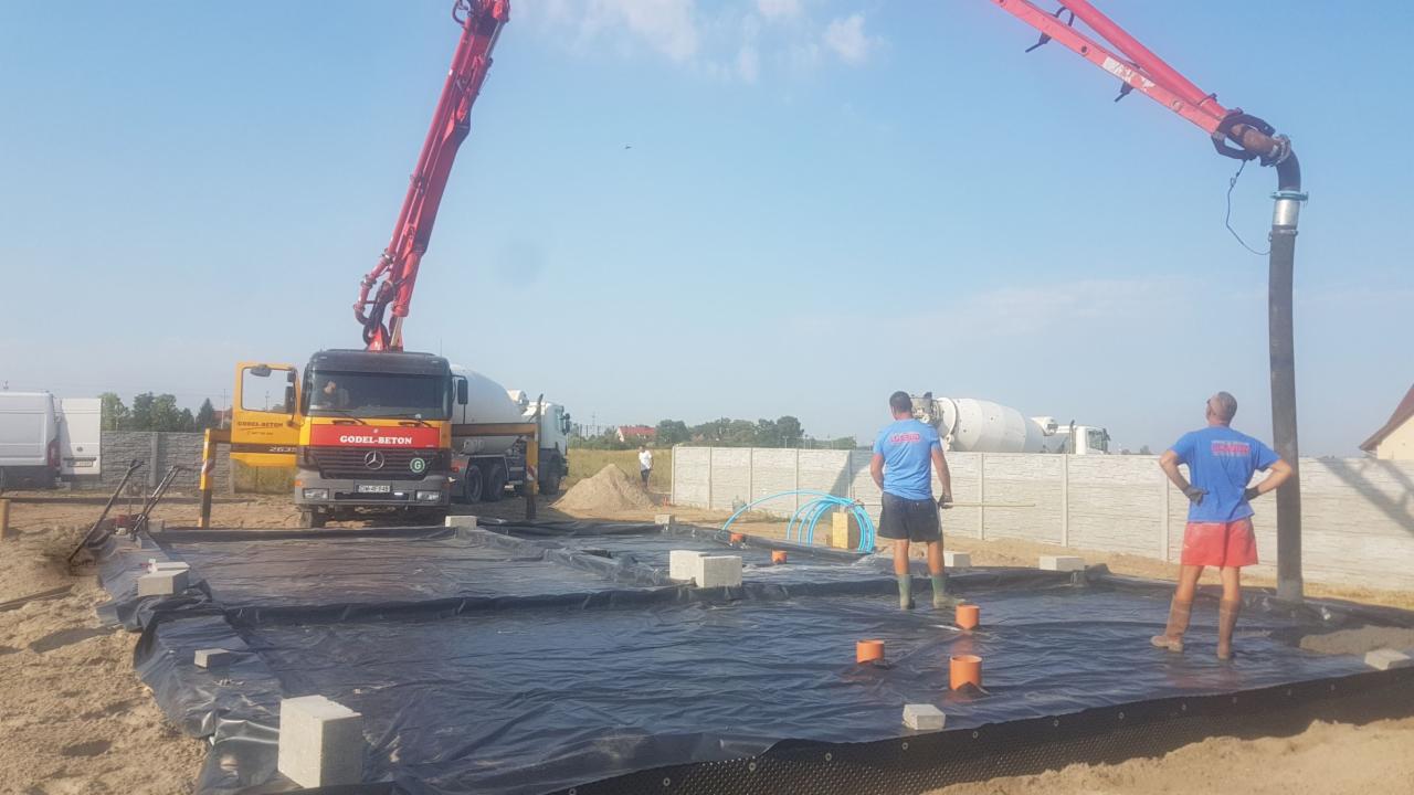 Przygotowanie podłoża pod fundamenty: rozłożona folia hydroizolacyjna, betonowe bloczki, rury kanalizacyjne, w tle betonomieszarka z pompą do betonu Godel-Beton i dwóch pracowników w roboczych...
