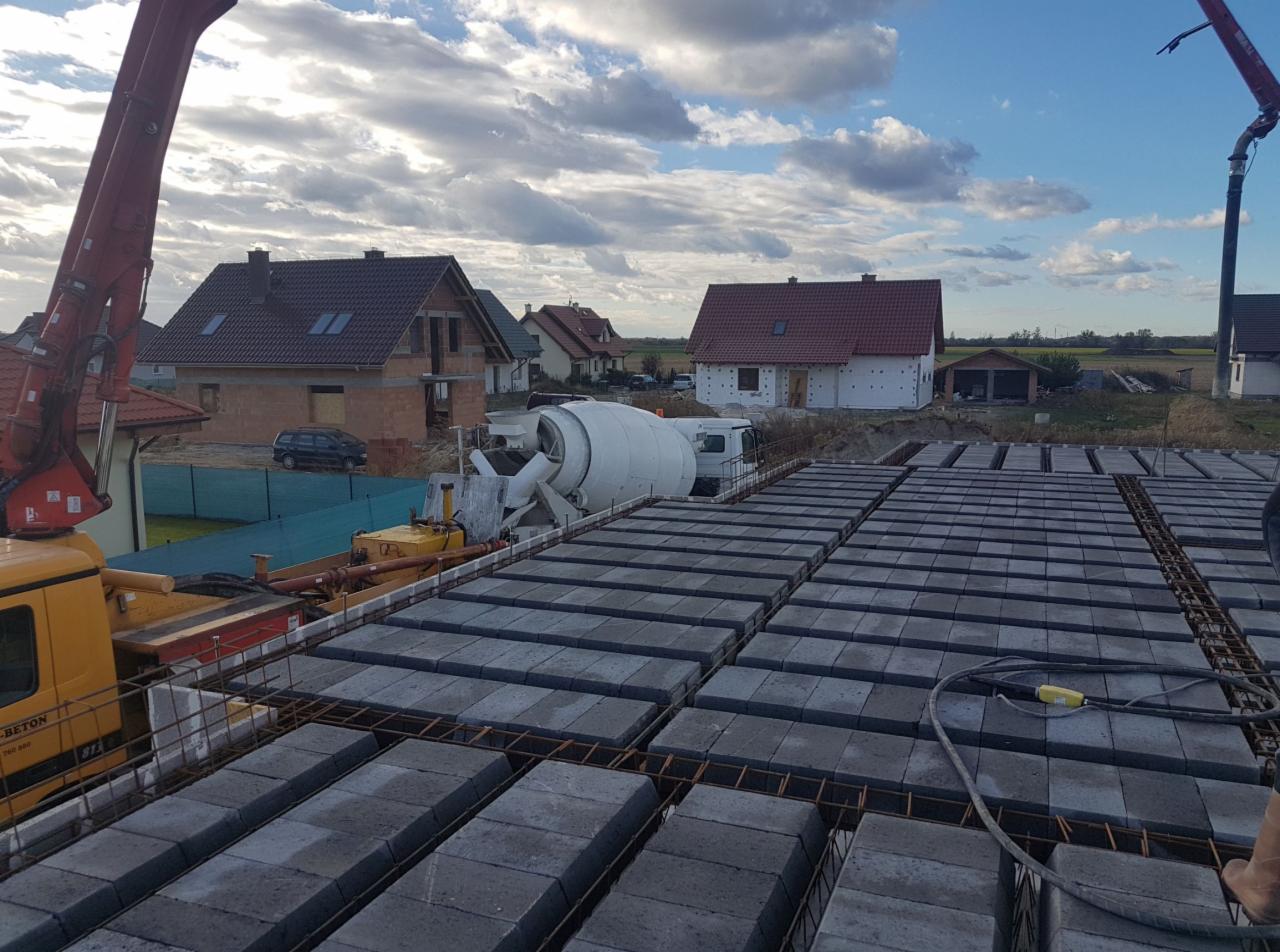 Wylany strop Teriva z widocznym żurawiem do betonu i betoniarką w tle, na działce budowlanej z domami w różnym stanie ukończenia.