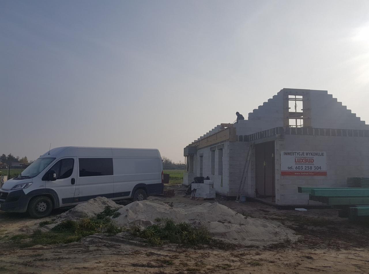 Budowa domu z białego bloczka: stan surowy otwarty, widoczny pracownik na dachu, biały samochód dostawczy firmy, sterta piasku na pierwszym planie, tablica informacyjna z nazwą firmy budowlanej...