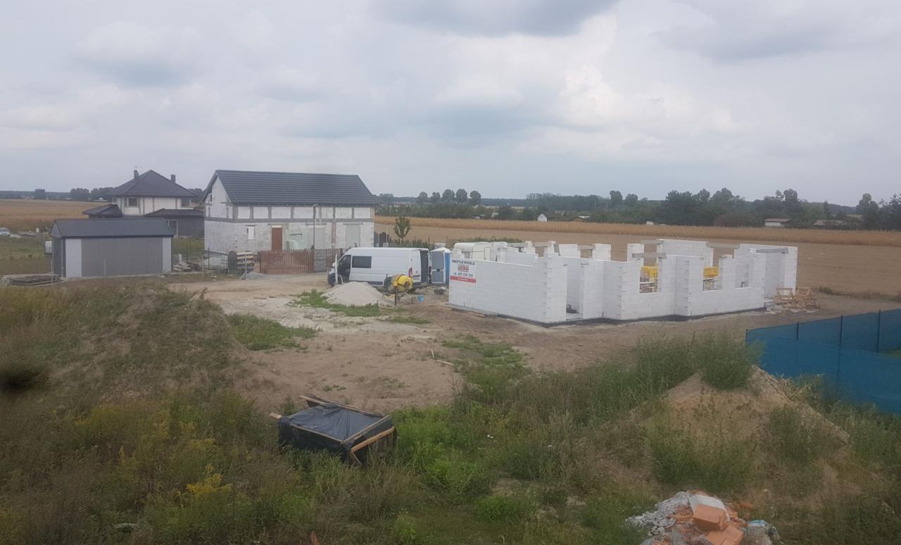 Budowa domu z bloczków betonu komórkowego w trakcie wznoszenia ścian, widoczny mikser do zaprawy, teren budowy z dwoma budynkami w tle i polem uprawnym.