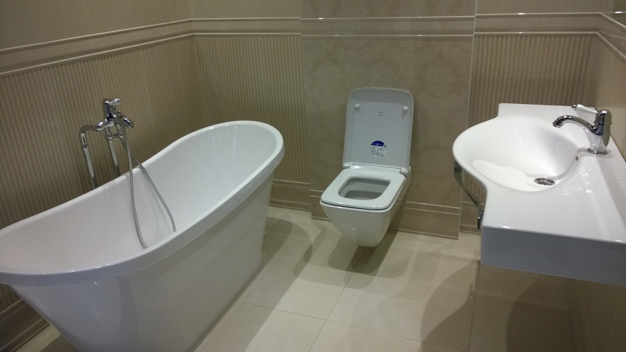 Biała, wolnostojąca wanna z chromowaną armaturą, wisząca toaleta z deską oraz umywalka nablatowa w beżowej łazience z płytkami imitującymi tapetę.