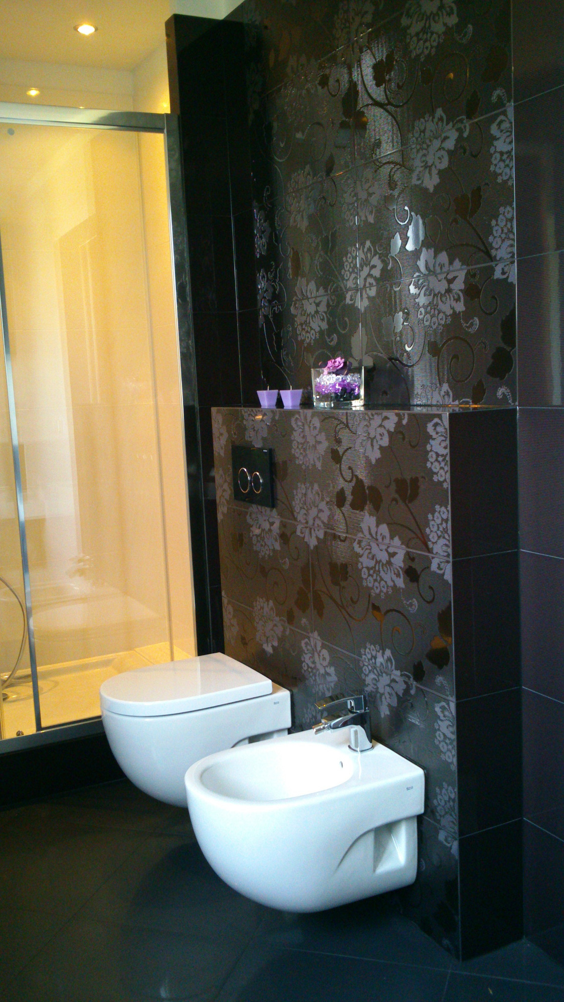 Nowoczesna łazienka z wiszącą toaletą i bidetem, ciemne płytki z kwiatowym wzorem, szklana kabina prysznicowa w tle, fioletowe dekoracje na półce nad toaletą.