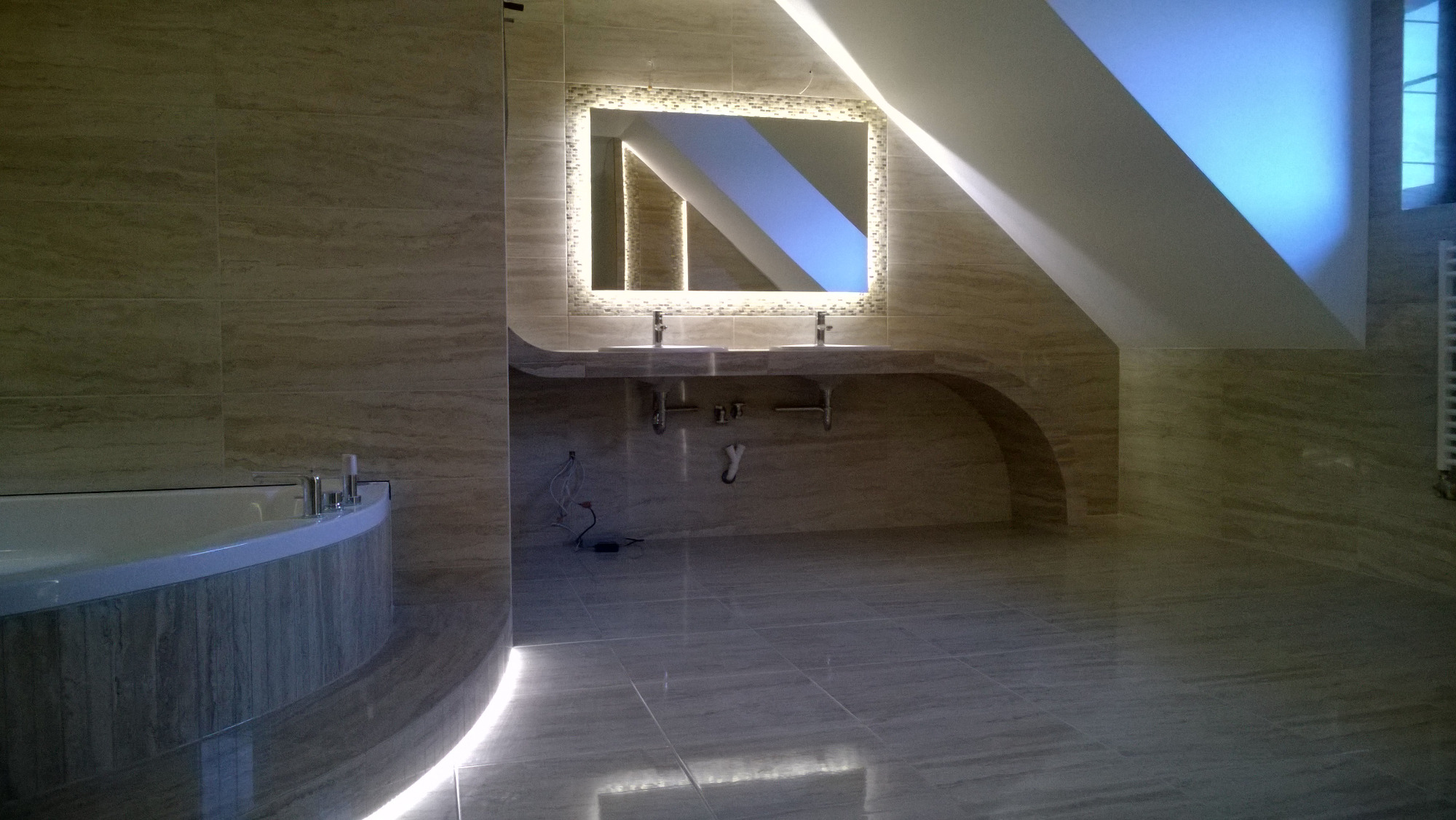 Przestronna łazienka na poddaszu z beżowymi płytkami imitującymi trawertyn, podświetlanym lustrem mozaikowym nad umywalkami i owalną wanną z podświetleniem LED.